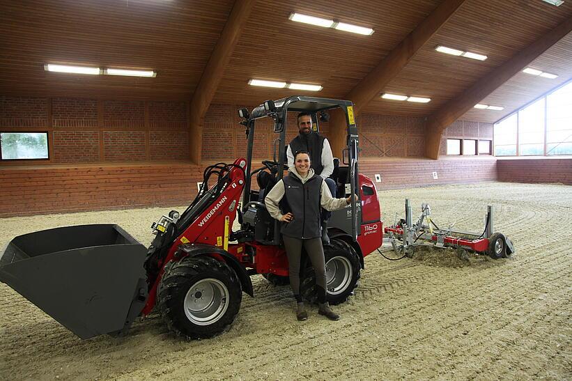 Weidemann Hoftrac 1160e mit einem Mann und einer Frau in einer Reithalle mit Leichtgutschaufel und Reitplatz- und Bahnenplaner